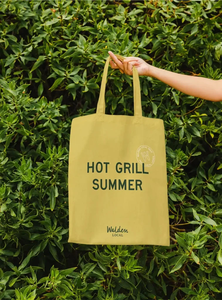 CPG branding agency work for Walden Local tote bag design with "hot grill summer" slogan held by a person outdoors.