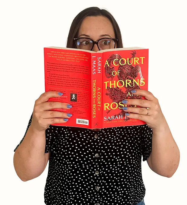 A woman in glasses and a black polka dot shirt reads A Court of Thorns and Roses by Sarah J. Maas, holding the book open in front of her face.