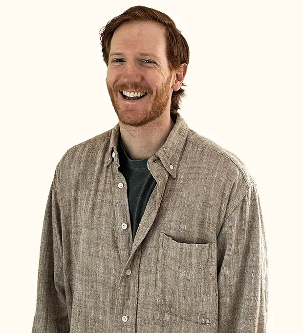 A man with reddish hair and a beard smiles, wearing a beige button-up shirt over a dark t-shirt, standing against a plain light background.