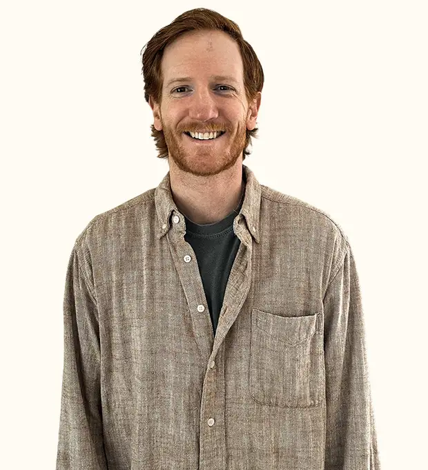 A smiling man with red hair and a beard, wearing a gray t-shirt under a beige button-up shirt, stands against a plain light background.