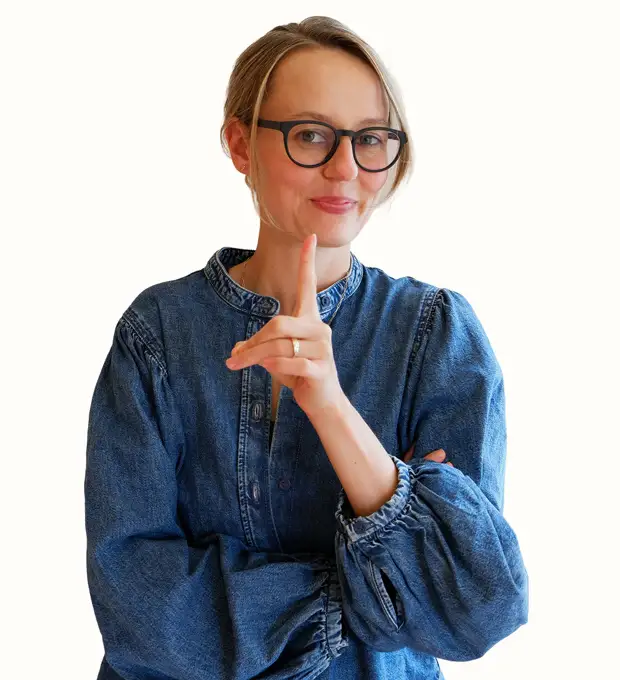 A person wearing glasses and a blue denim shirt holds a finger to their lips, signaling to be quiet, with a slight smile and their other arm crossed.
