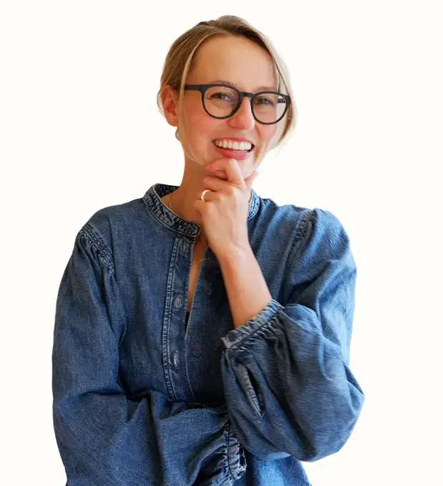 A smiling woman wearing glasses and a denim shirt, standing with her hand thoughtfully touching her chin against a plain white background.