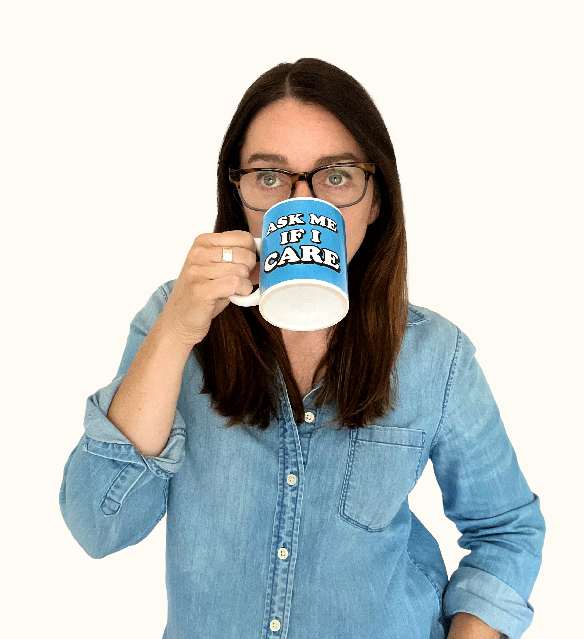 Woman with long brown hair and glasses wears a denim shirt, holding a mug that says ASK ME IF I CARE while drinking from it, against a plain white background.