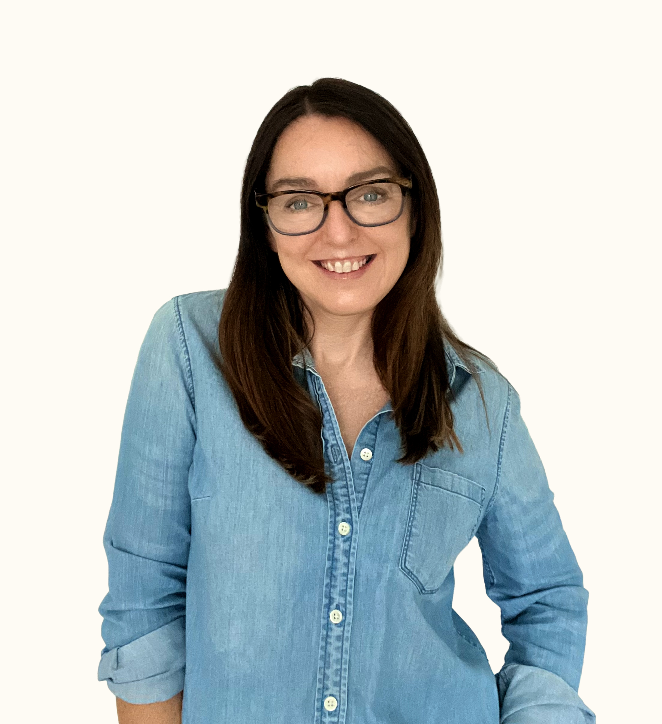 Woman with long brown hair and glasses, smiling, wearing a light blue denim shirt with rolled-up sleeves, standing against a plain white background.