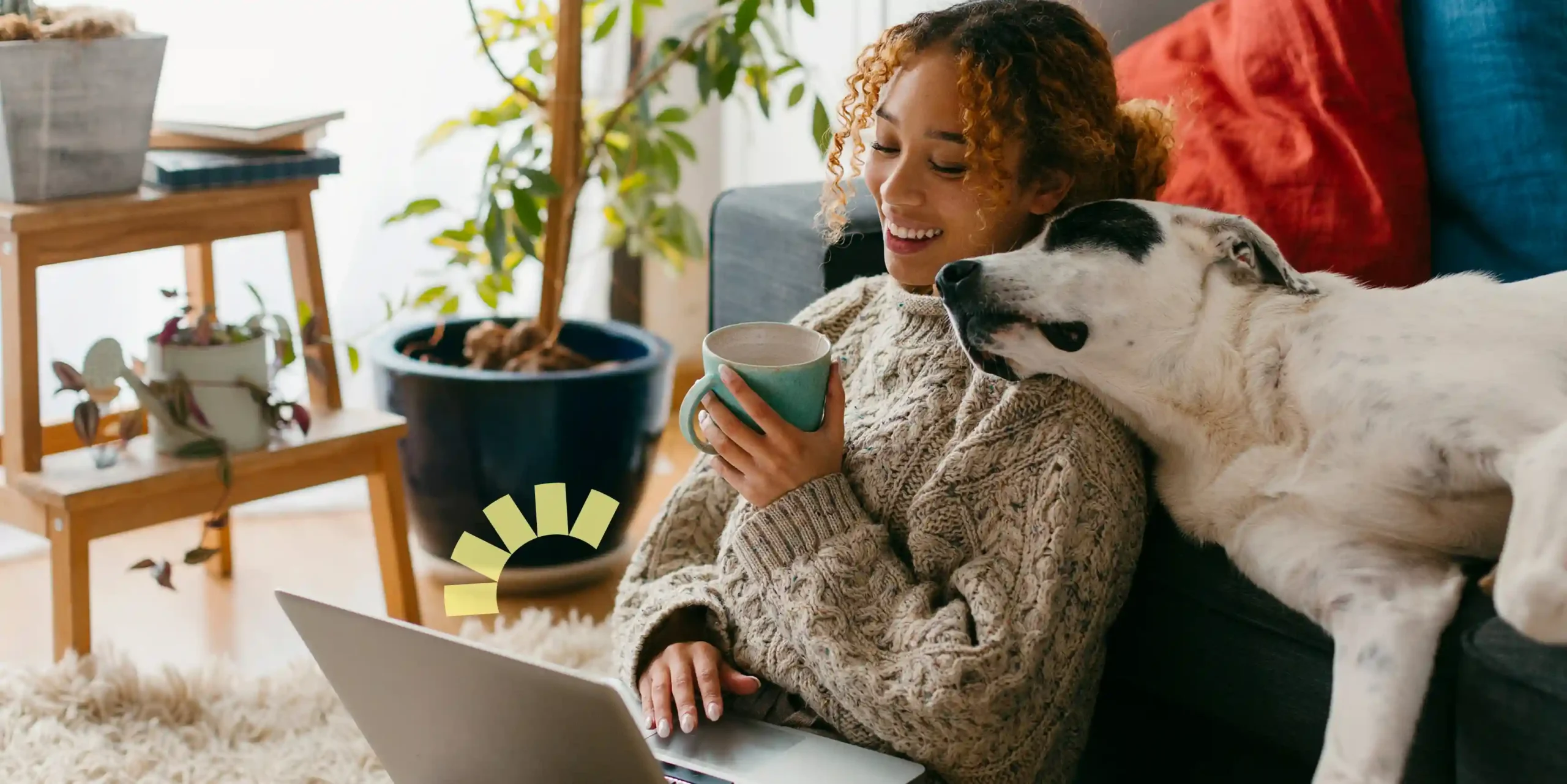 A woman sits on the floor with a laptop, smiling and holding a mug, while a black and white dog rests its head on her shoulder. A plant and couch are in the background.