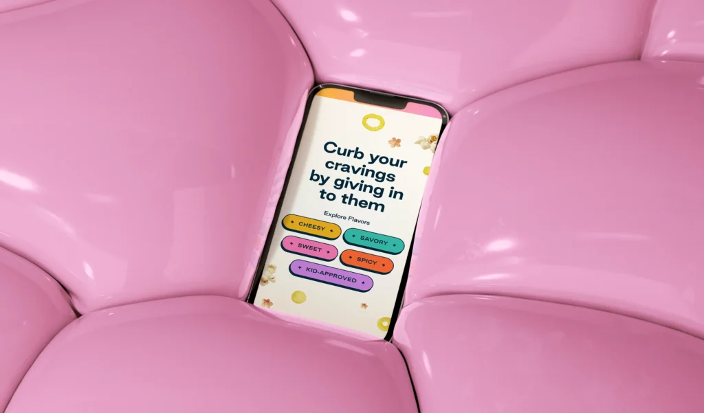 A smartphone displaying a cravings app rests on shiny, soft, pink cushioned surfaces. The app screen invites users to explore flavors like sweet, savory, spicy, and kid-approved.