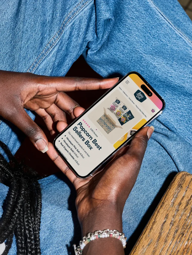 A person holding a smartphone with a “Popcorn Best Sellers Box” product page open; they are wearing jeans and a beaded bracelet.