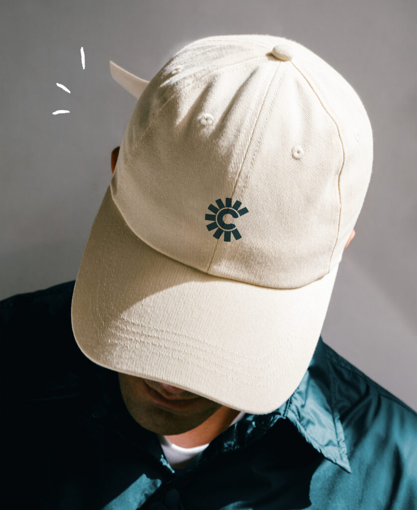 A person wearing a cream-colored baseball cap with a circular, stylized “C” logo, looking down, in a dark collared shirt.