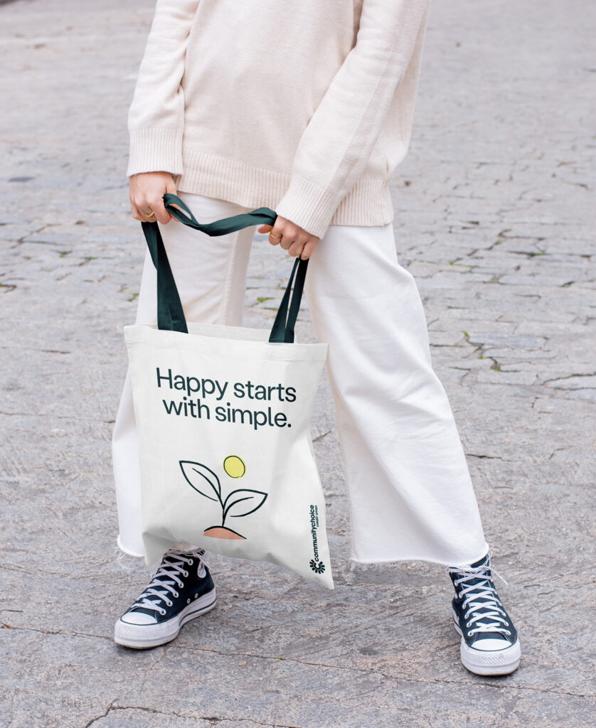 A person in white clothes and black sneakers holds a tote bag that says Happy starts with simple. with a plant and sun illustration printed on it.