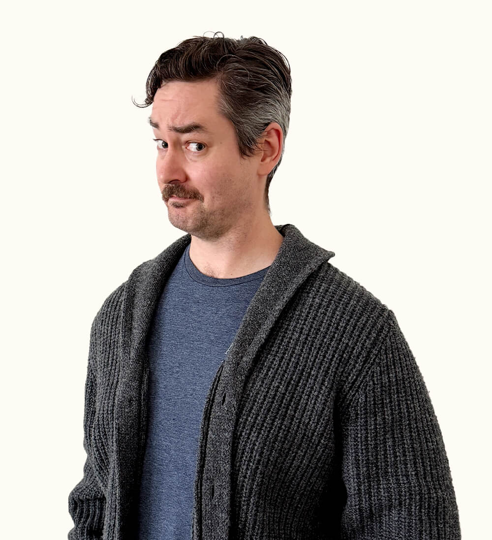 A man with short dark hair and a mustache, wearing a blue shirt and gray cardigan, looks to the side with a raised eyebrow, expressing skepticism.