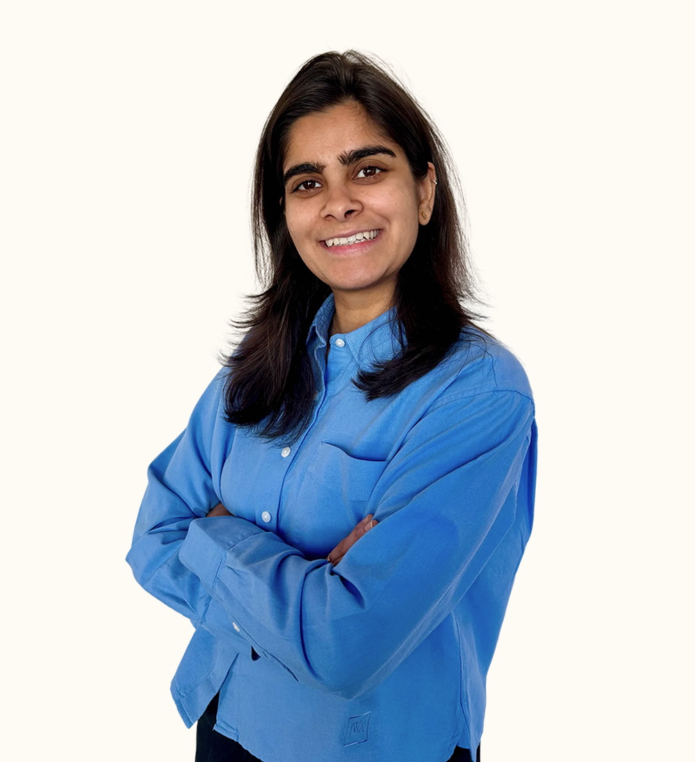 A person with long dark hair, wearing a blue shirt, smiles with their arms crossed against a plain white background.