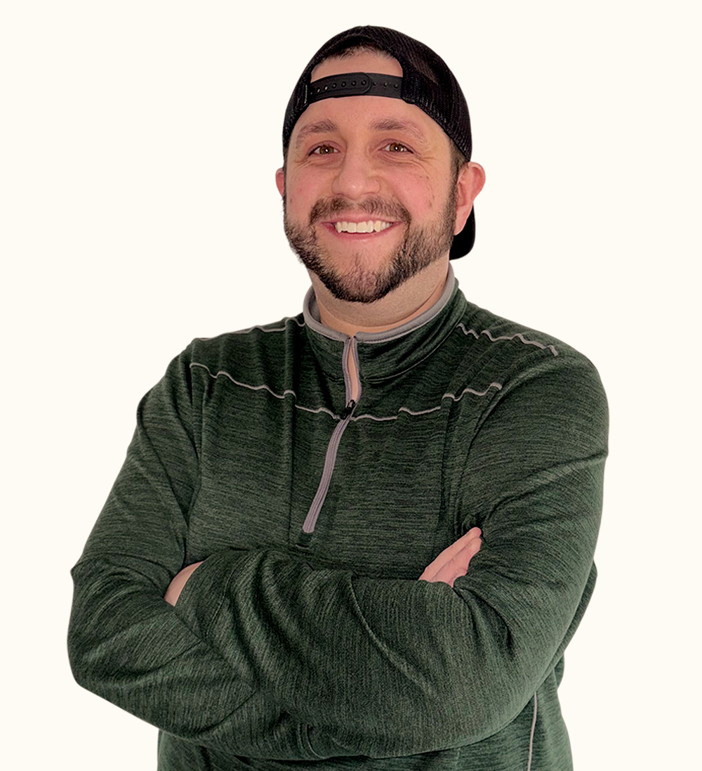 A man in a dark green pullover and black backwards cap smiles with arms crossed, standing against a light background.