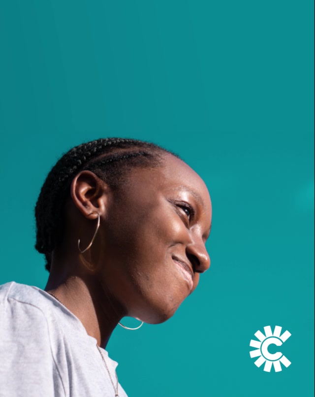 A person with braided hair and hoop earrings smiles, looking up against a bright teal background. A white circular logo appears in the lower right corner.