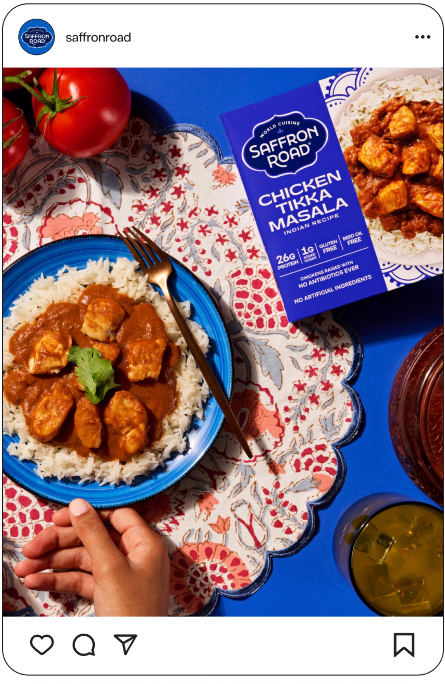 A hand holds a fork over a plate of chicken tikka masala on rice, next to a Saffron Road meal box, fresh tomatoes, a drink, and a doily on a blue table.