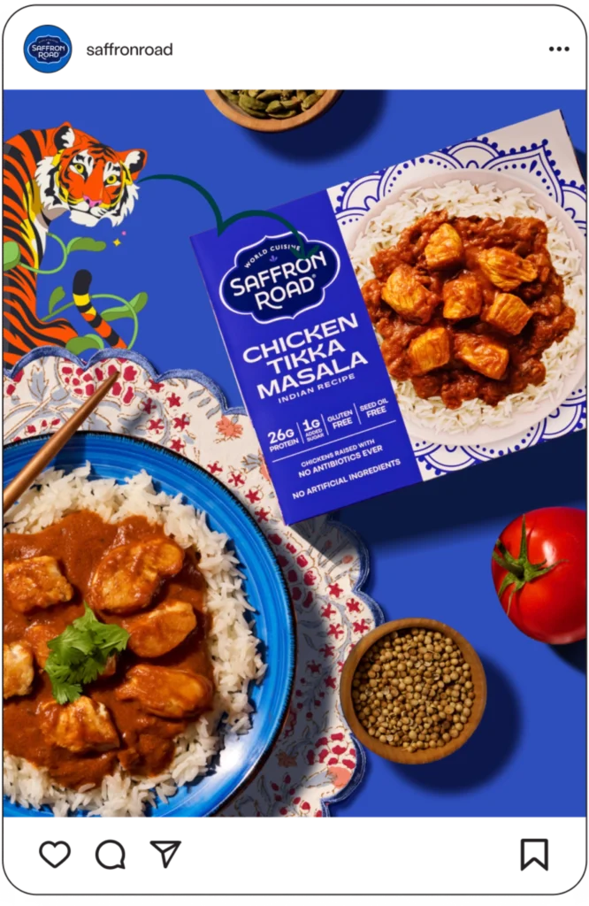 A plate of chicken tikka masala with rice sits beside its Saffron Road packaging, on a blue table with spices, a tomato, and a tiger illustration in the background.