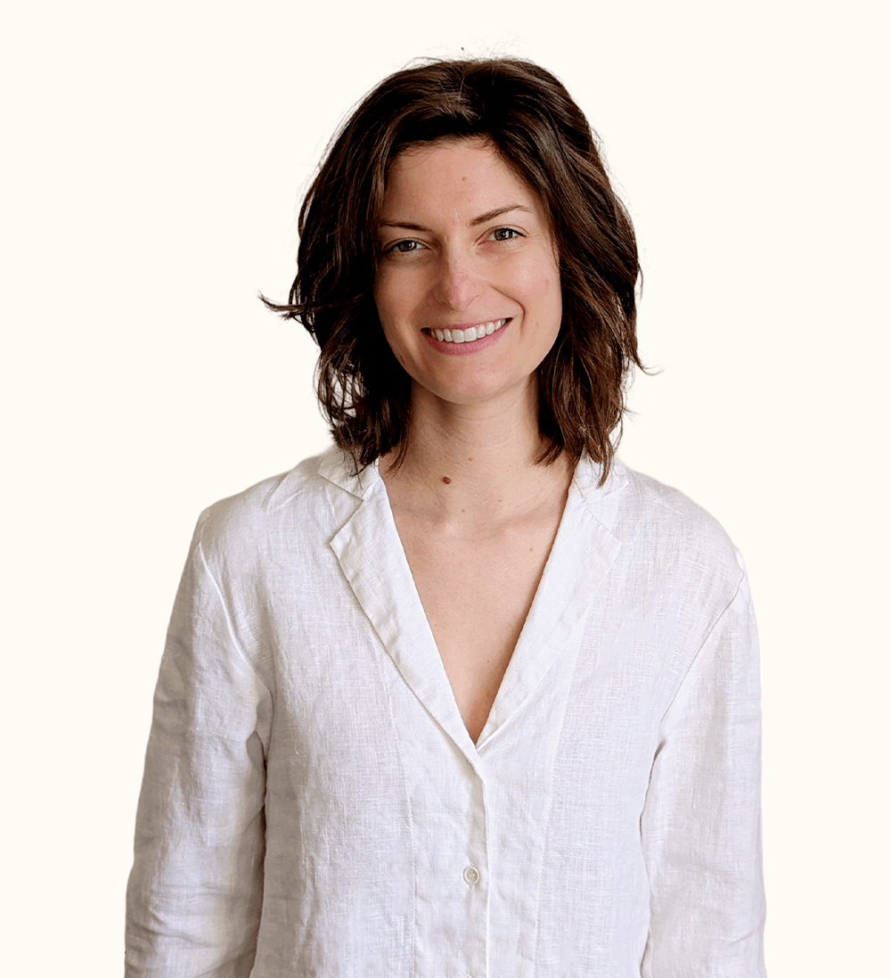 A woman with wavy brown hair wearing a white linen shirt smiles while standing against a plain, light background.