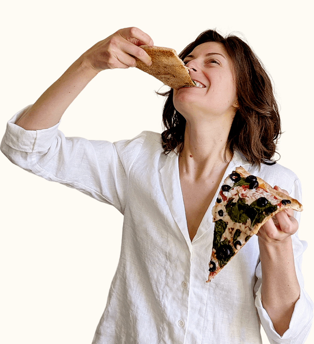 A woman in a white shirt smiles and eats a slice of pizza, while holding another slice in her other hand.