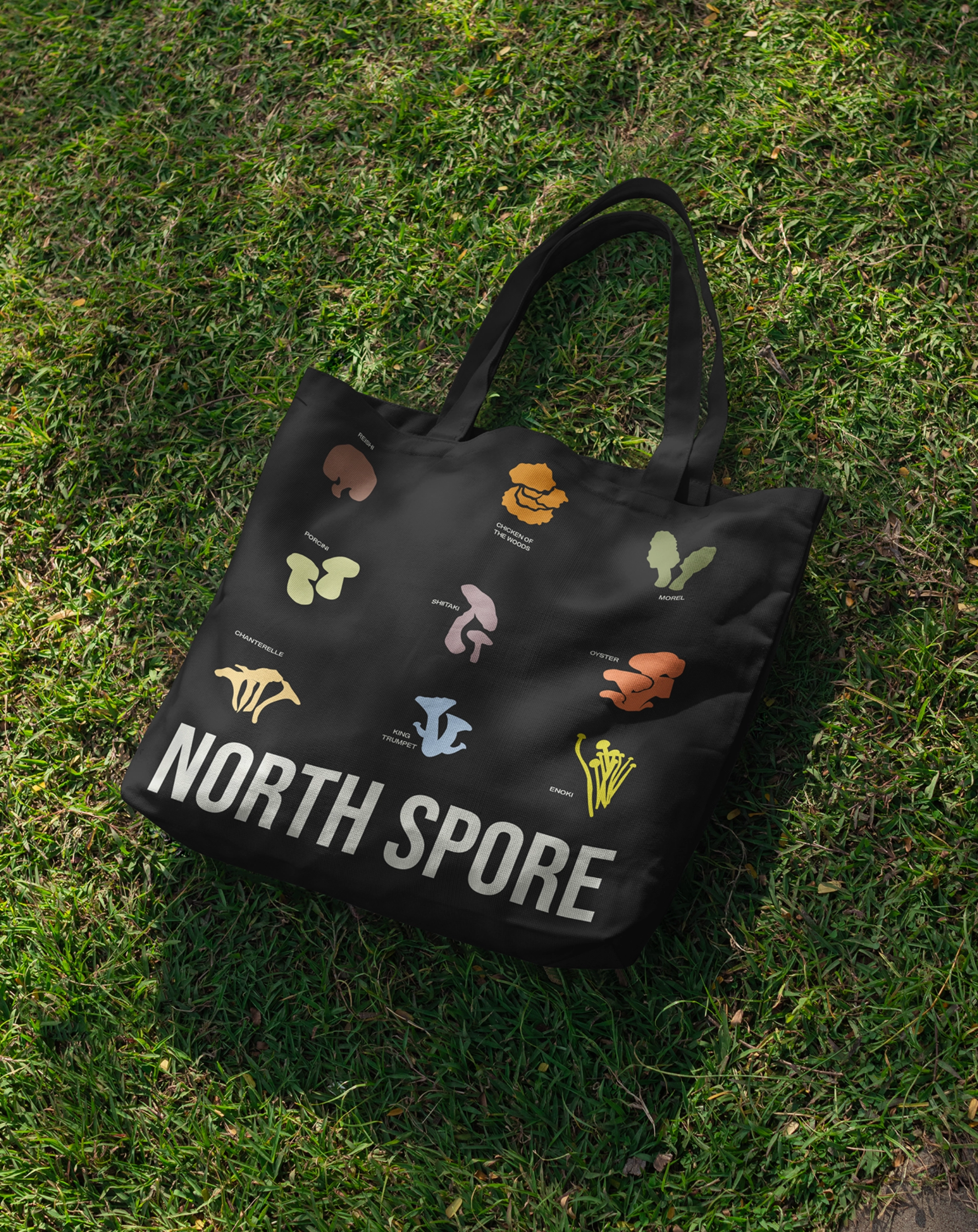 A black tote bag with mushroom illustrations and names, labeled NORTH SPORE, lying on green grass in sunlight.