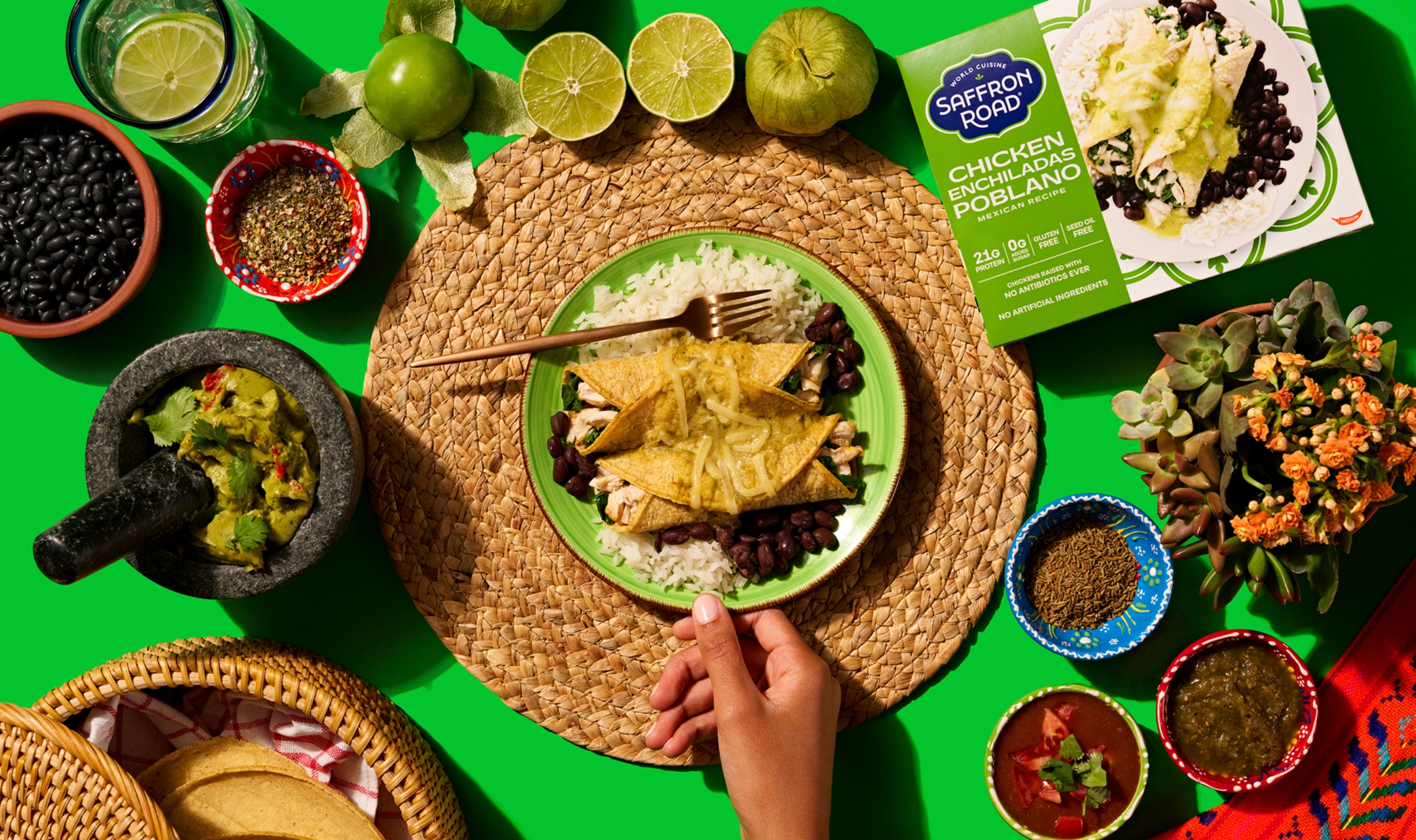 A hand holds a fork over a plate of enchiladas with rice and black beans, surrounded by Mexican ingredients, sauces, and a Saffron Road enchiladas package on a green background.