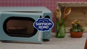 A blue microwave sits on a countertop beside a green jar with wooden utensils, a small plant, and a wooden box; Saffron Road logo is centered over the image.