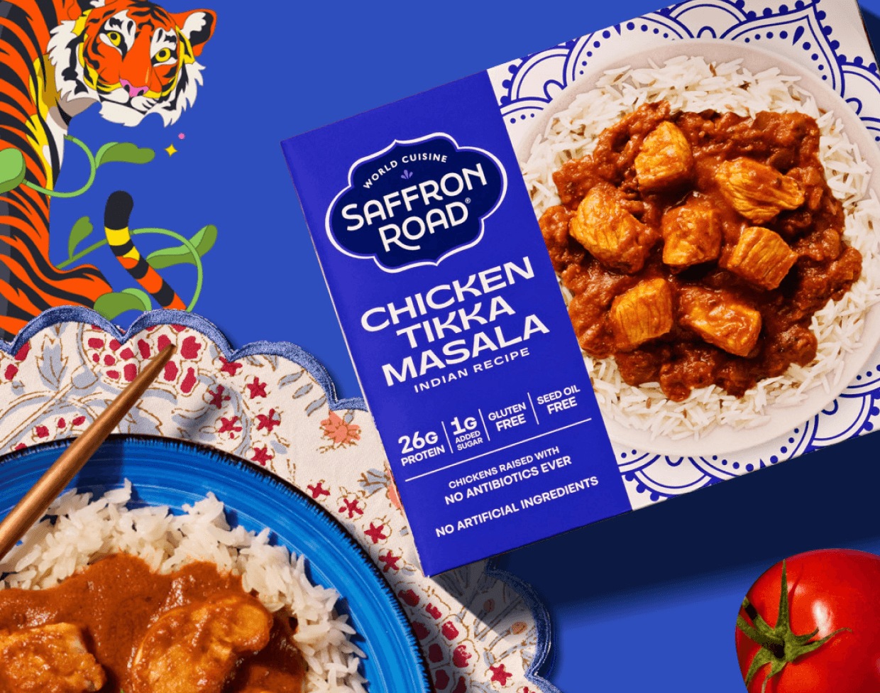A box of Saffron Road Chicken Tikka Masala sits on a plate of rice, with chopsticks and curry nearby, against a vibrant blue background with a tiger illustration.