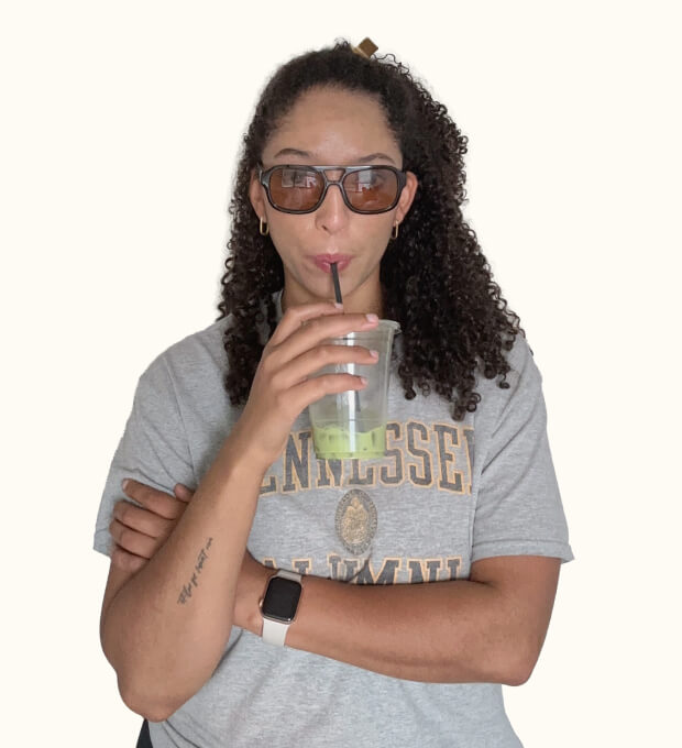 A person with curly hair, sunglasses, and a gray Tennessee Alumni shirt drinks a green beverage with a straw, arms crossed, and wearing a smartwatch.