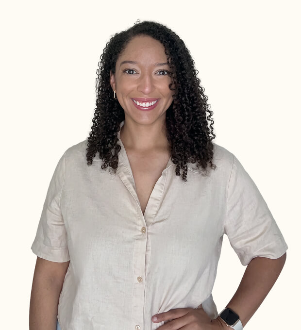 Smiling woman with curly hair, wearing a beige button-up shirt and a smartwatch, standing against a plain light background.