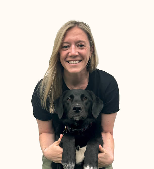 A smiling woman with long blonde hair holds a black dog with white paws. They are both facing the camera against a plain white background.