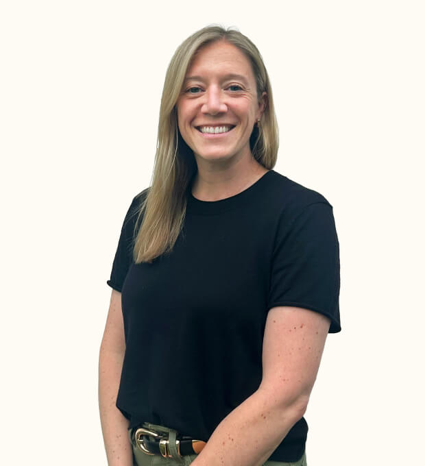 Smiling person with straight, light brown hair wears a black t-shirt and olive-green pants, standing against a plain, light background.