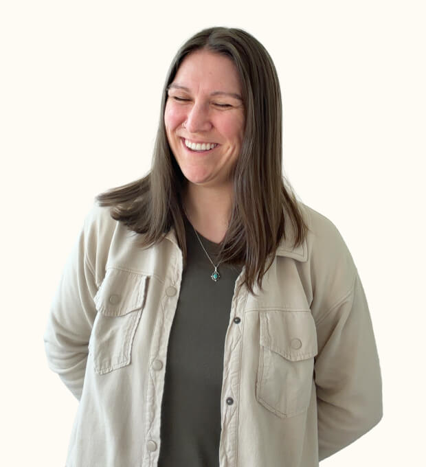 Woman with straight brown hair smiles with eyes closed, wearing a beige button-up shirt over a dark top, standing against a plain light background.