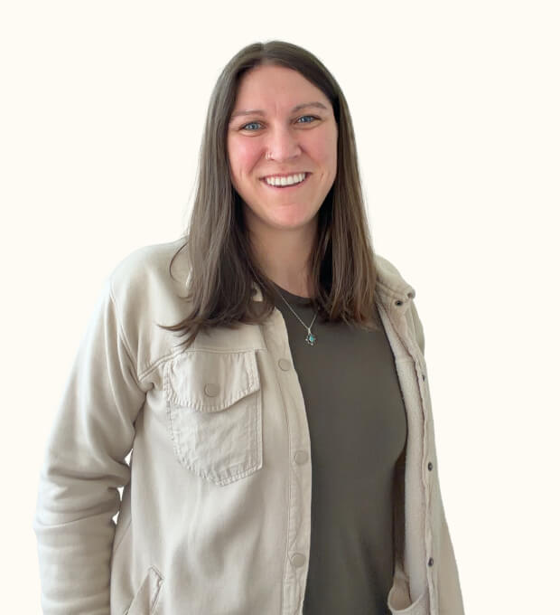 Woman with long brown hair, smiling, wearing a light beige jacket over a dark shirt, standing against a plain light background.