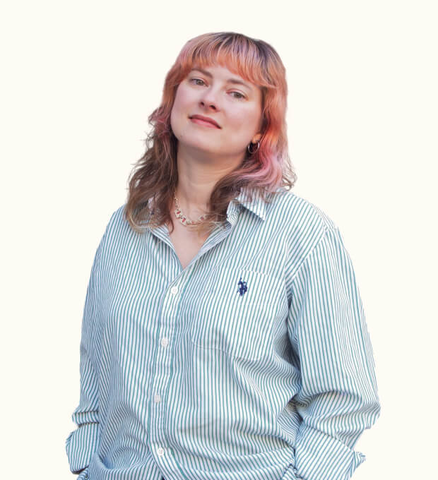 Person with wavy, shoulder-length hair wearing a striped button-up shirt, standing against a plain light background and looking at the camera with a slight smile.