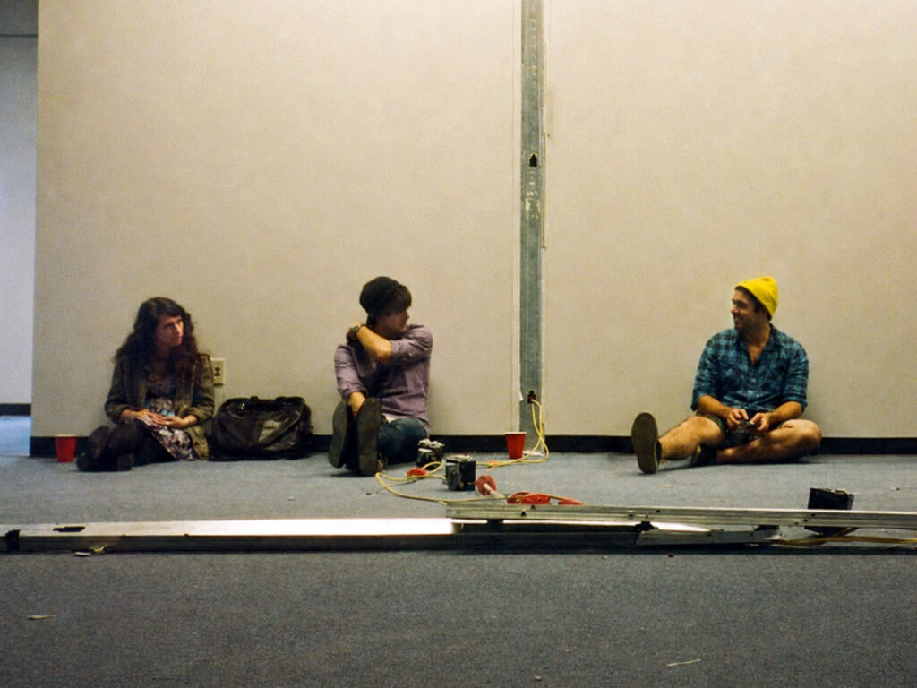 Three people sit on a carpeted floor against a bare wall, talking. There are bags, cups, and miscellaneous items scattered near them.