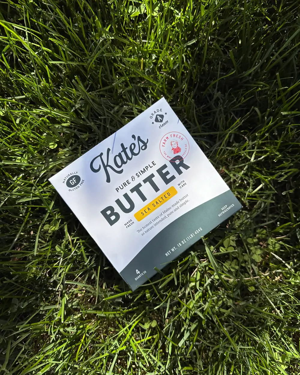 A package of Kates Sea Salted Butter rests on green grass in bright sunlight. The butter package has white and green colors and visible text.