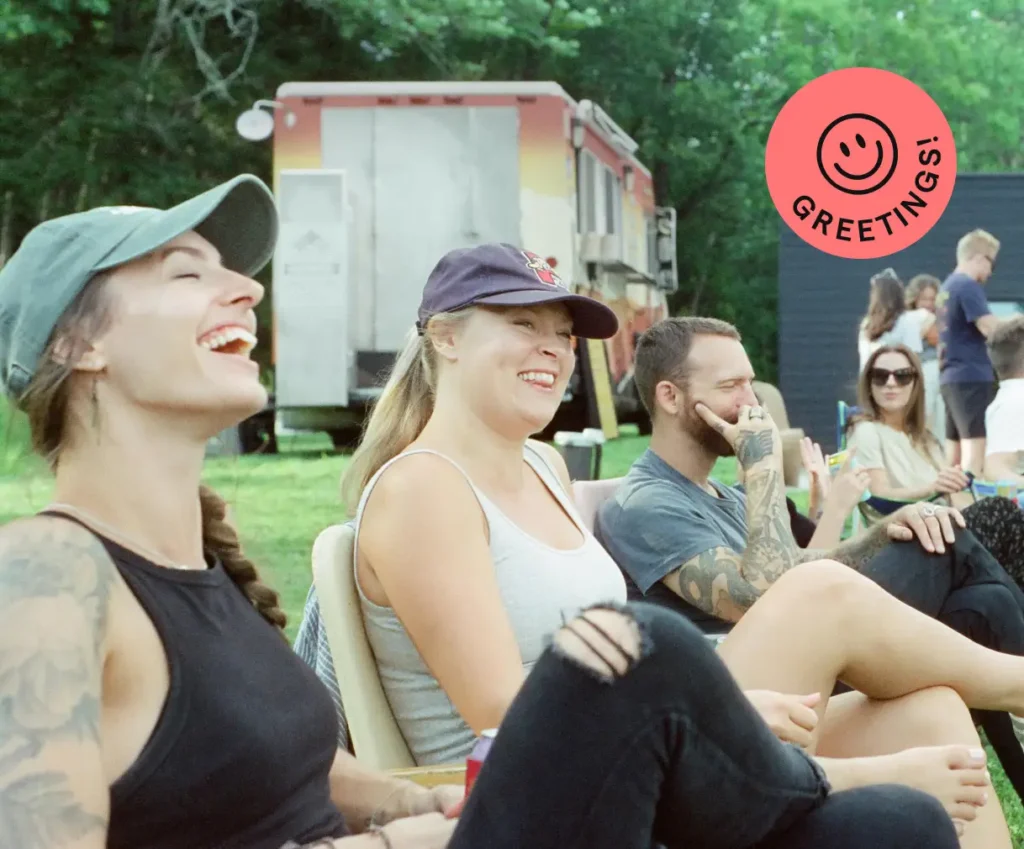 Three people sit outside on chairs, laughing and smiling. A Greetings! sticker with a smiley face is in the corner. A food truck and more people are in the background.