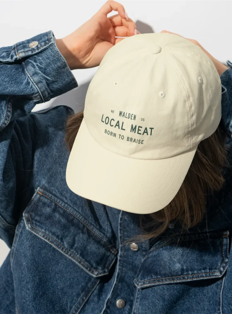 Person in a denim jacket adjusts a beige baseball cap reading WALDEN LOCAL MEAT BORN TO BRAISE in green text. Face is mostly hidden by the cap.